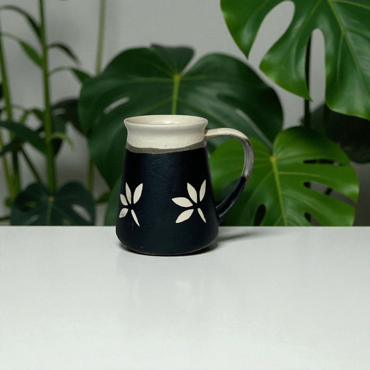 Black mug with white floral designs on a white surface with green leaves in the background