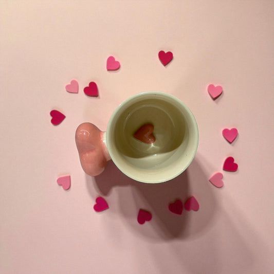 Hand holding a small cup with a heart-shaped object on a pink background with scattered hearts.