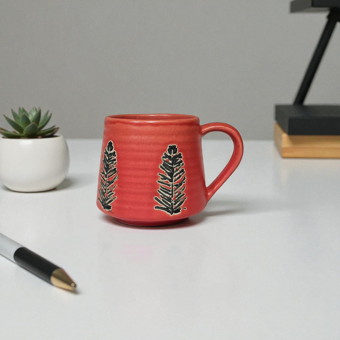 Red mug with black leaf design next to a small potted plant on a white surface.