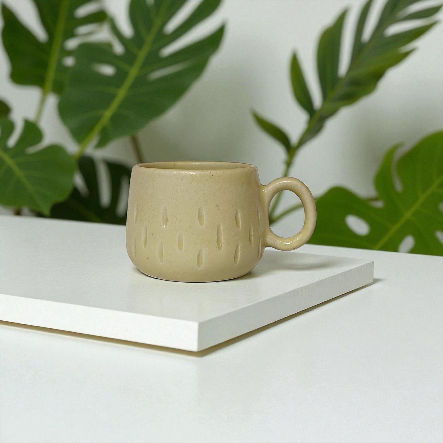Beige ceramic mug on a white surface with green leaves in the background