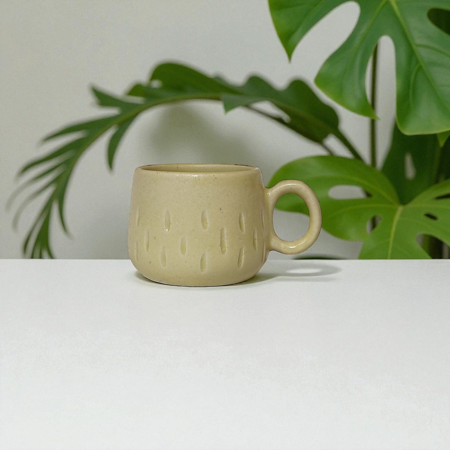 Beige ceramic mug on a white surface with green leaves in the background