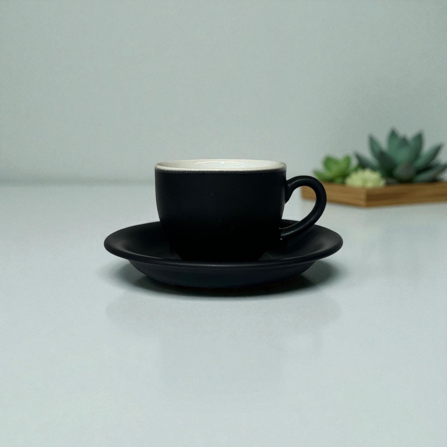 Black coffee cup and saucer on a light gray surface with a blurred plant in the background