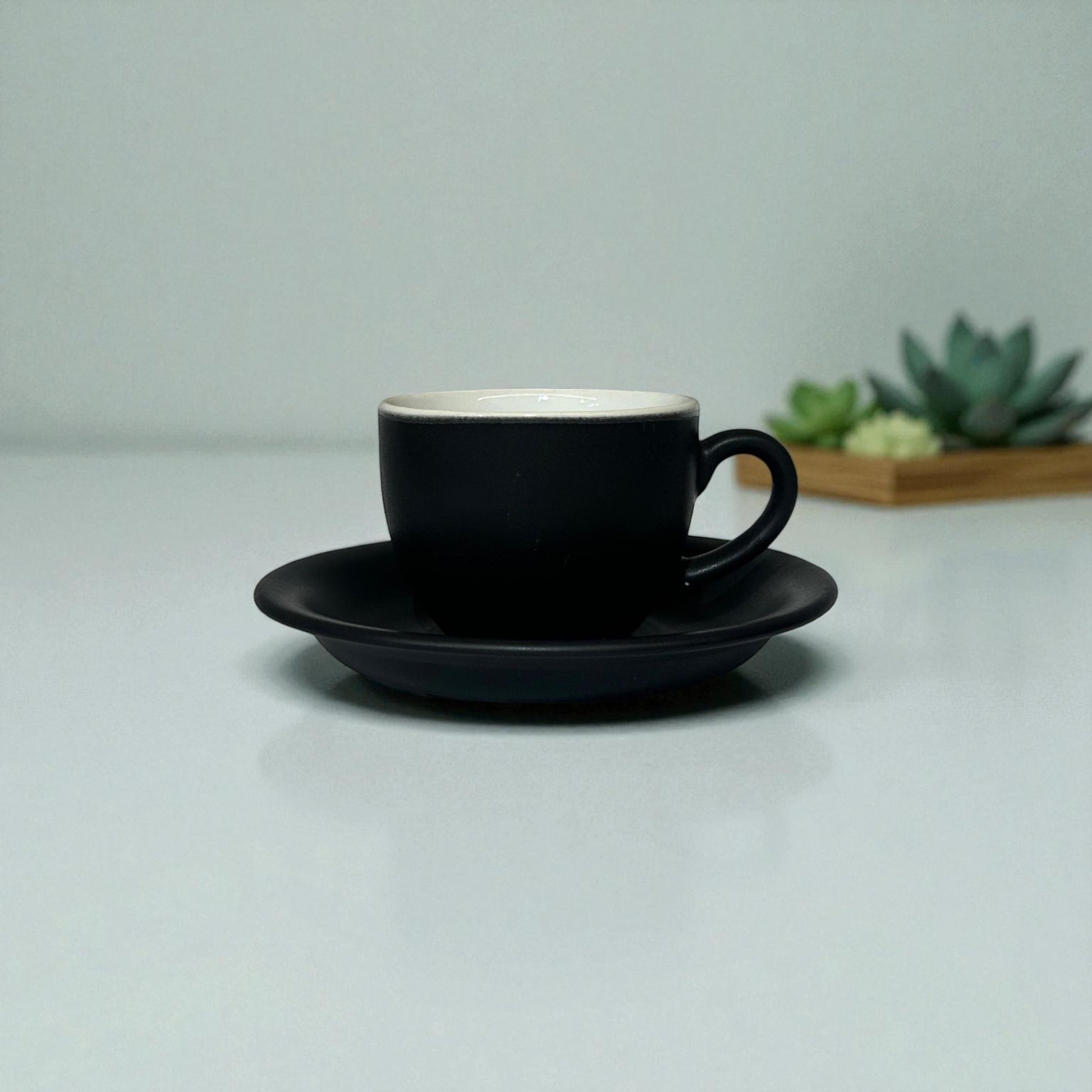 Black coffee cup and saucer on a light gray surface with a blurred plant in the background