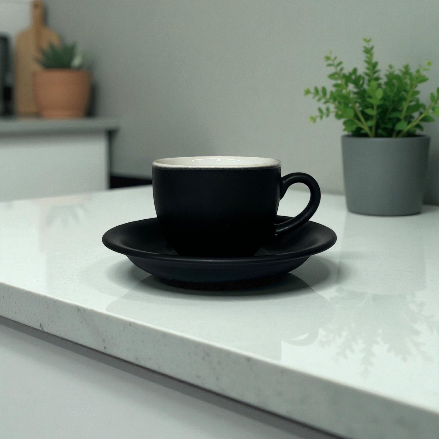 Black coffee cup and saucer on a white countertop with plants in the background