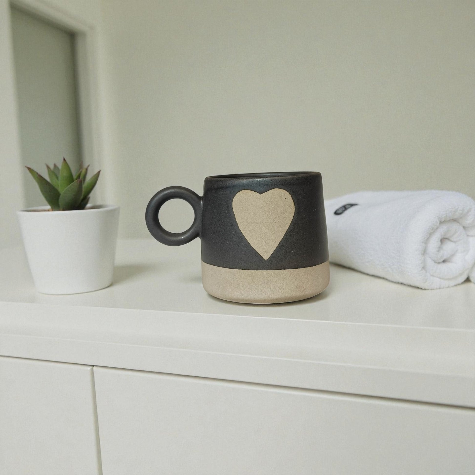 Black mug with a heart design on a white surface next to a plant and towel