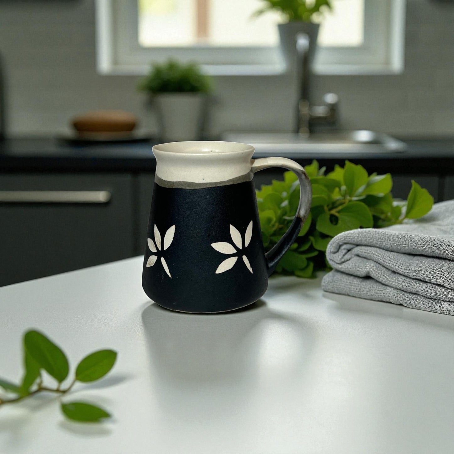 Black mug with white floral designs on a kitchen counter with plants and a towel.