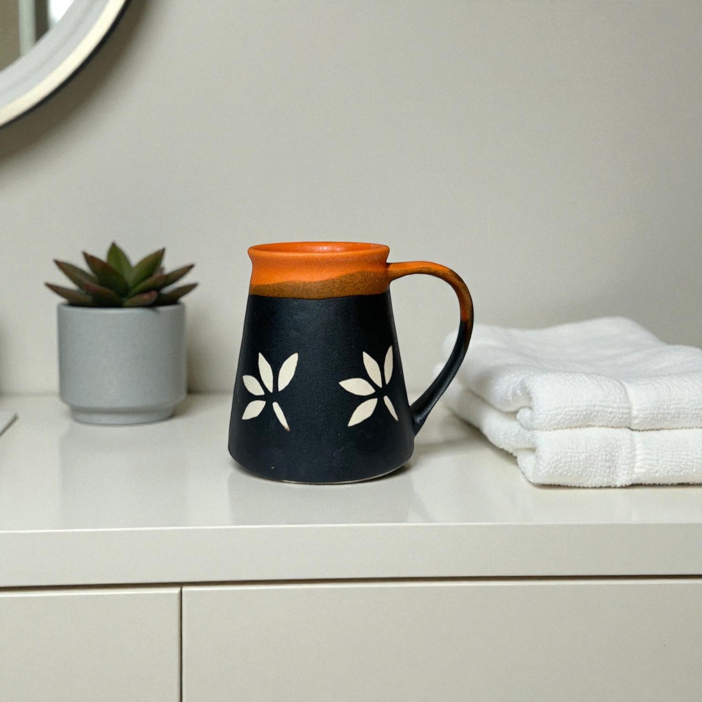 Black mug with white floral designs on a white surface next to a plant and folded towels.