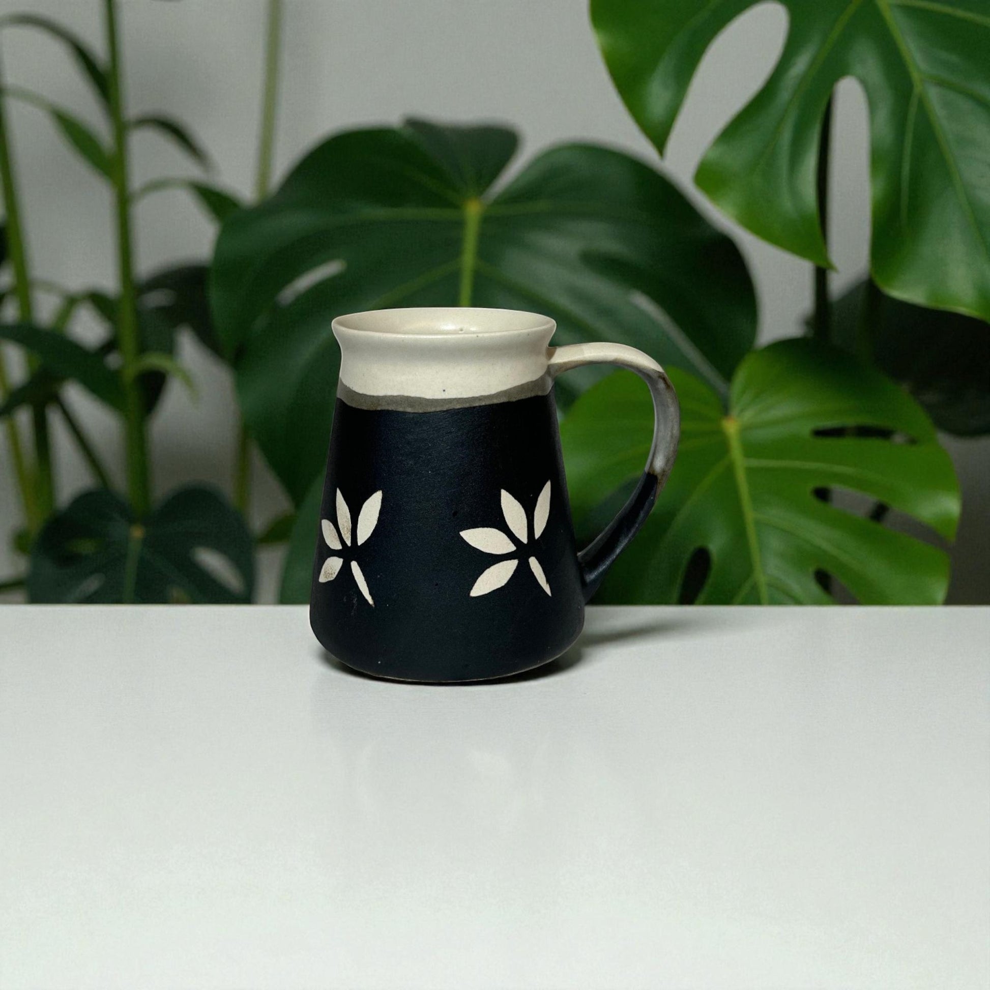 Black mug with white floral designs on a white surface with green leaves in the background