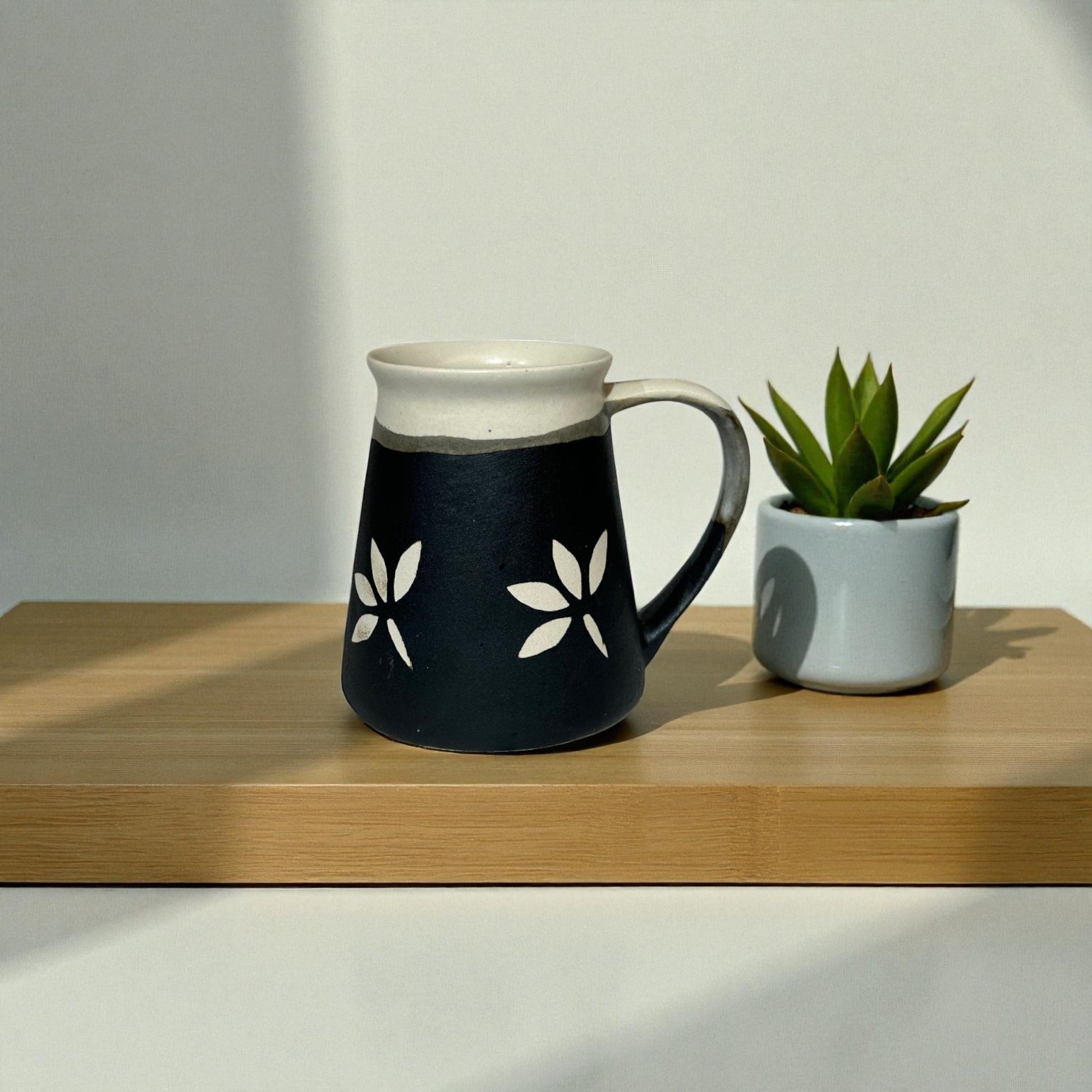 Black mug with white floral designs on a wooden surface next to a small potted plant.