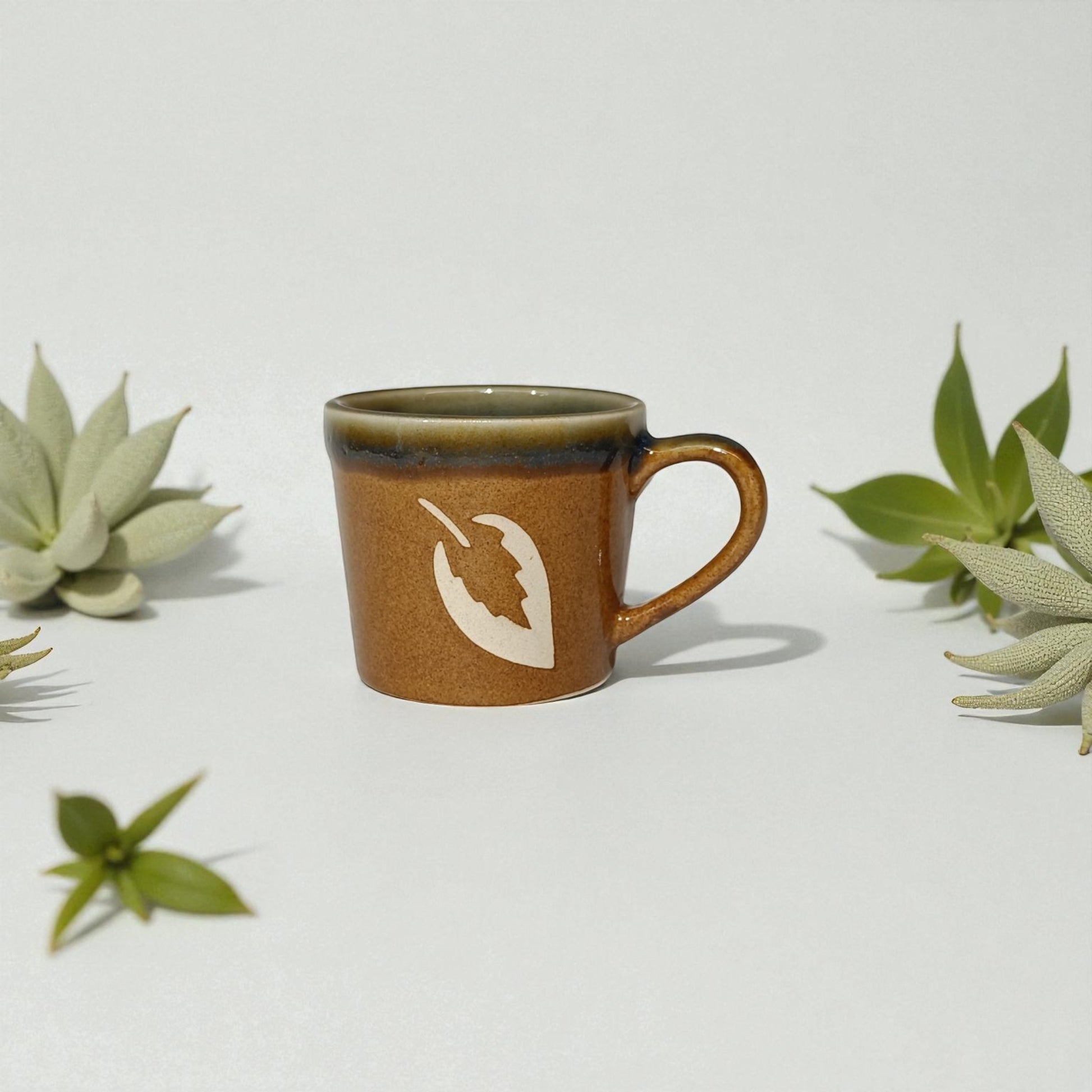 Brown ceramic mug with a white design on a light gray background with green plants.