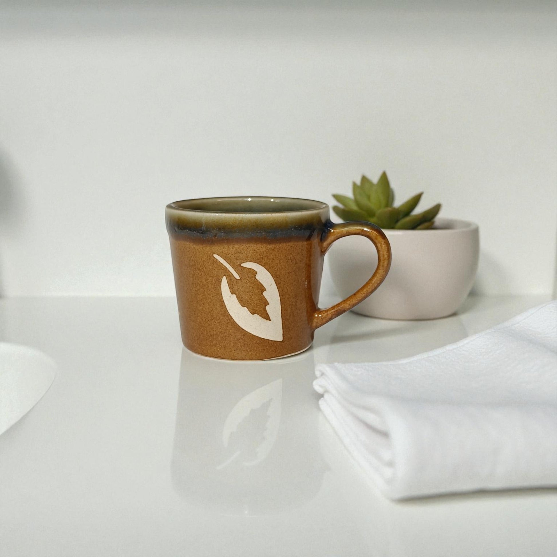 Brown ceramic mug with a white logo on a white surface with a plant in the background