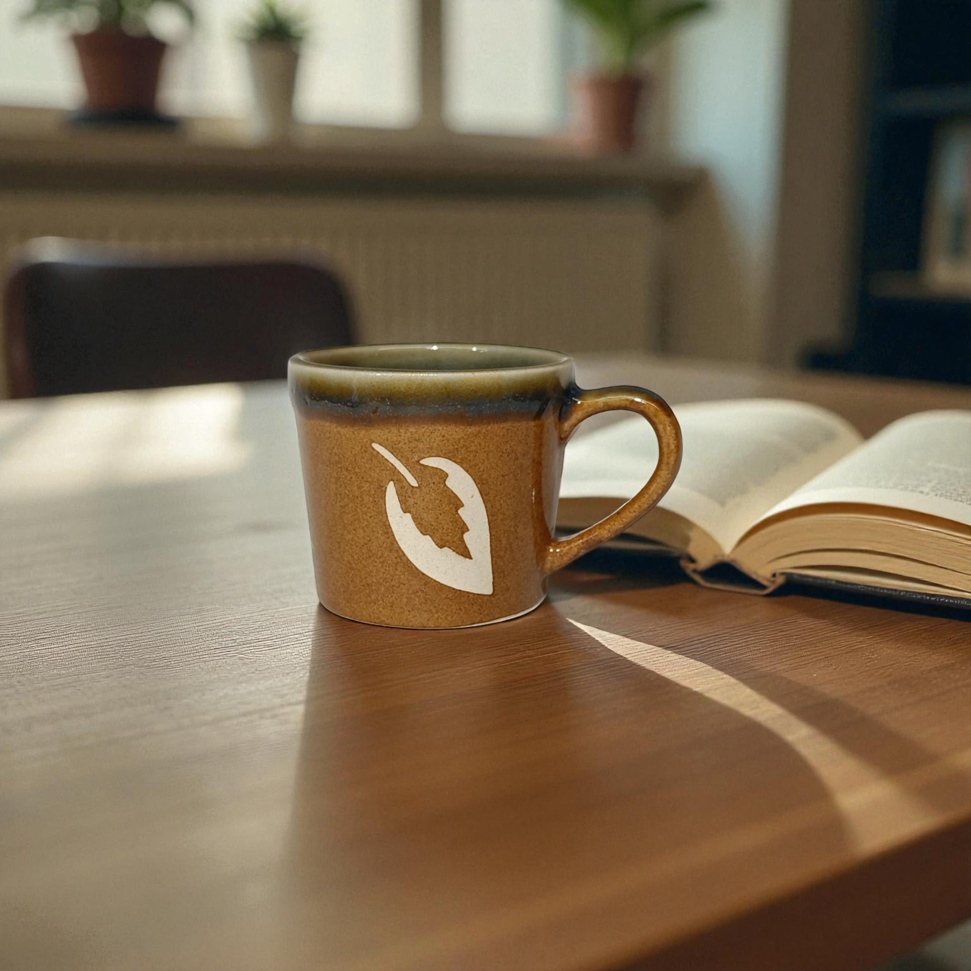 Brown mug with a white design on a wooden table next to an open book, with a blurred indoor background.
