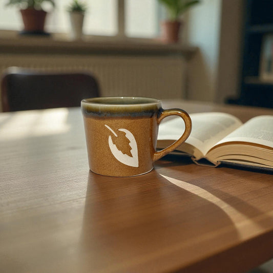 Brown mug with a white design on a wooden table next to an open book, with a blurred indoor background.