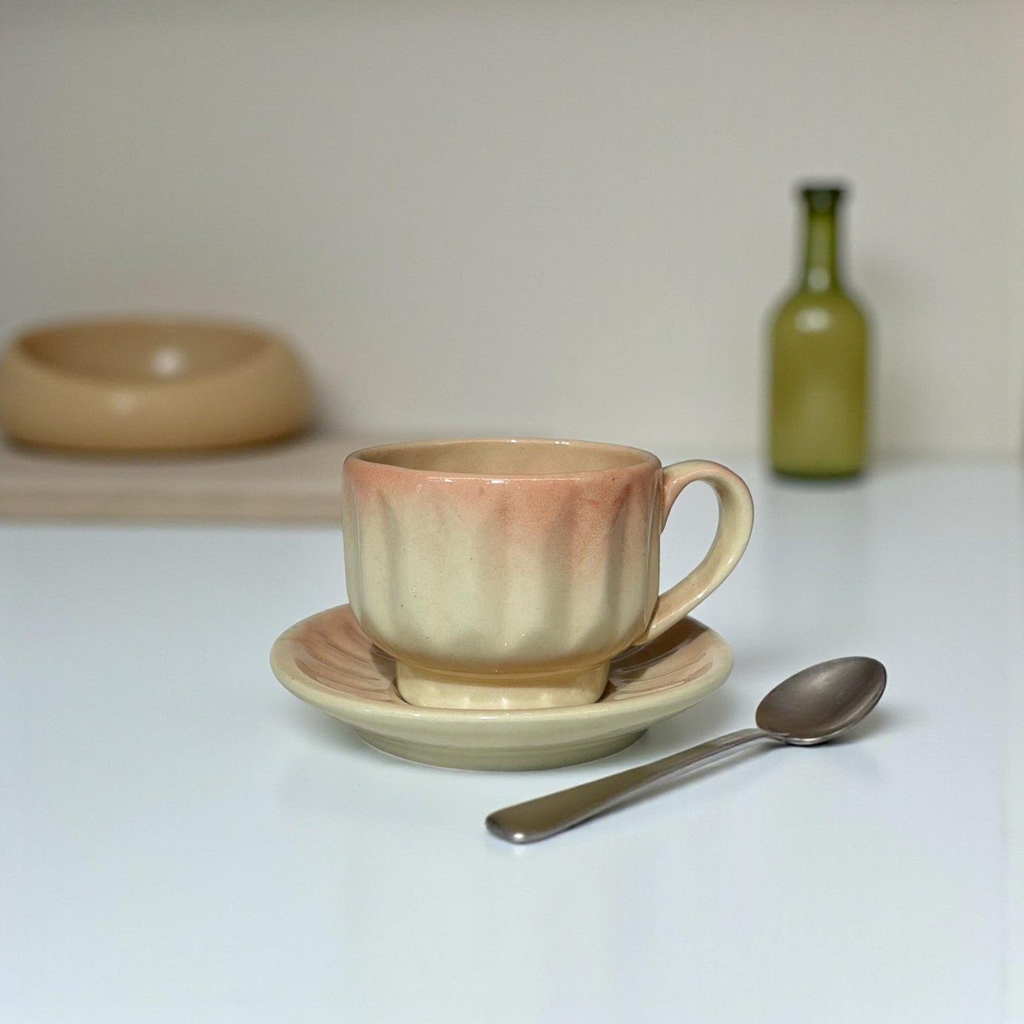 Ceramic cup and saucer with a spoon on a white surface, with a bottle in the background.