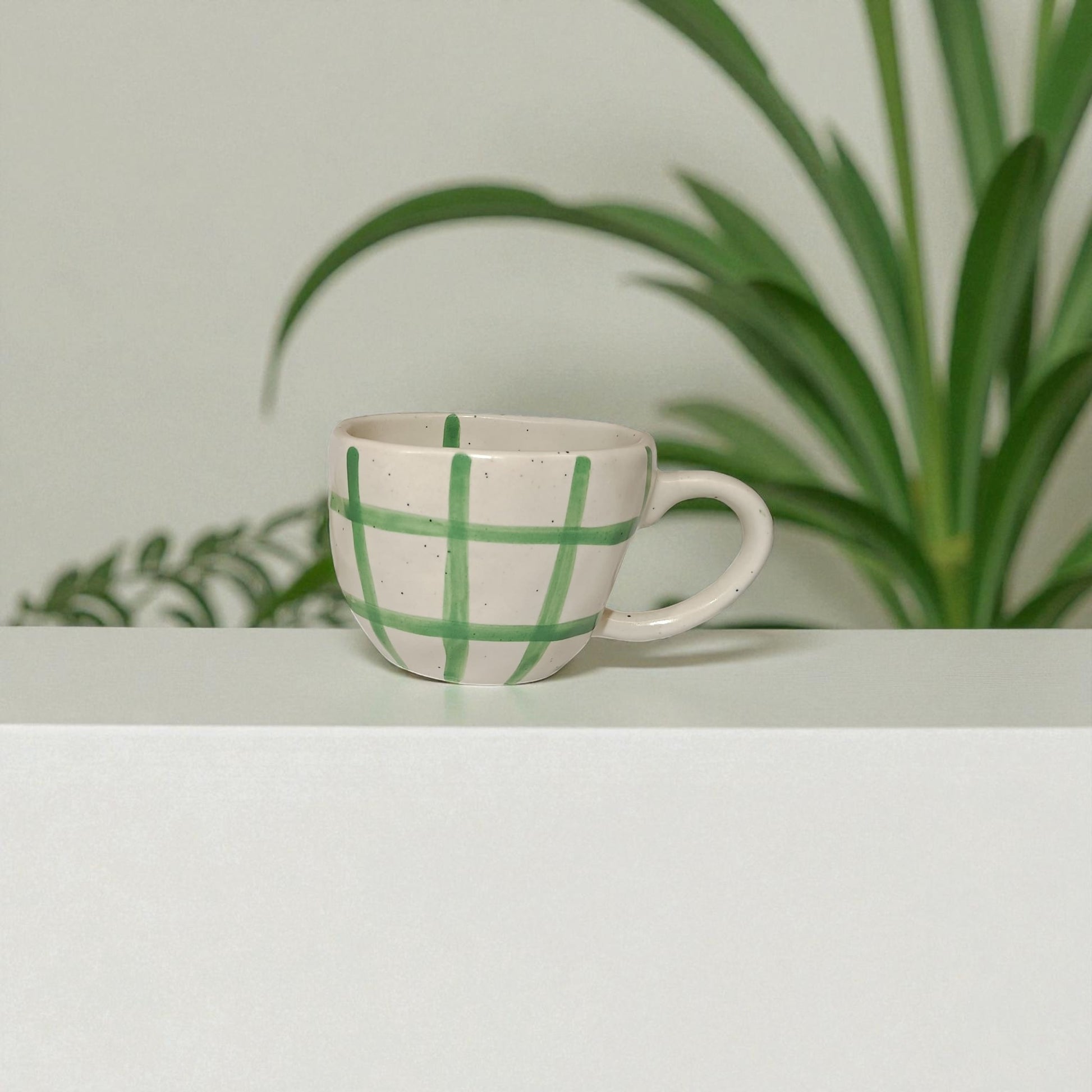 Ceramic cup with green checkered pattern on a light surface with plants in the background