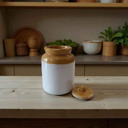 Ceramic jar with a brown lid on a wooden surface, with a shelf of kitchen items in the background.