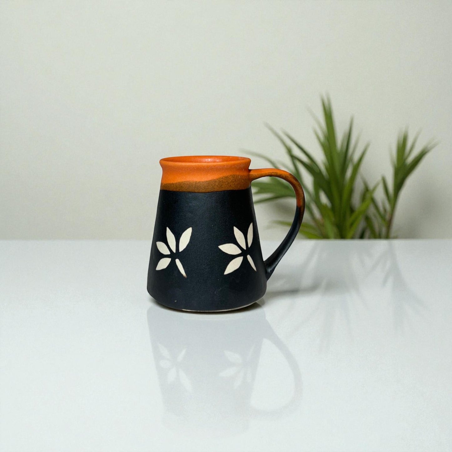 Ceramic mug with black body, orange rim, and white floral patterns on a white surface with a plant in the background.