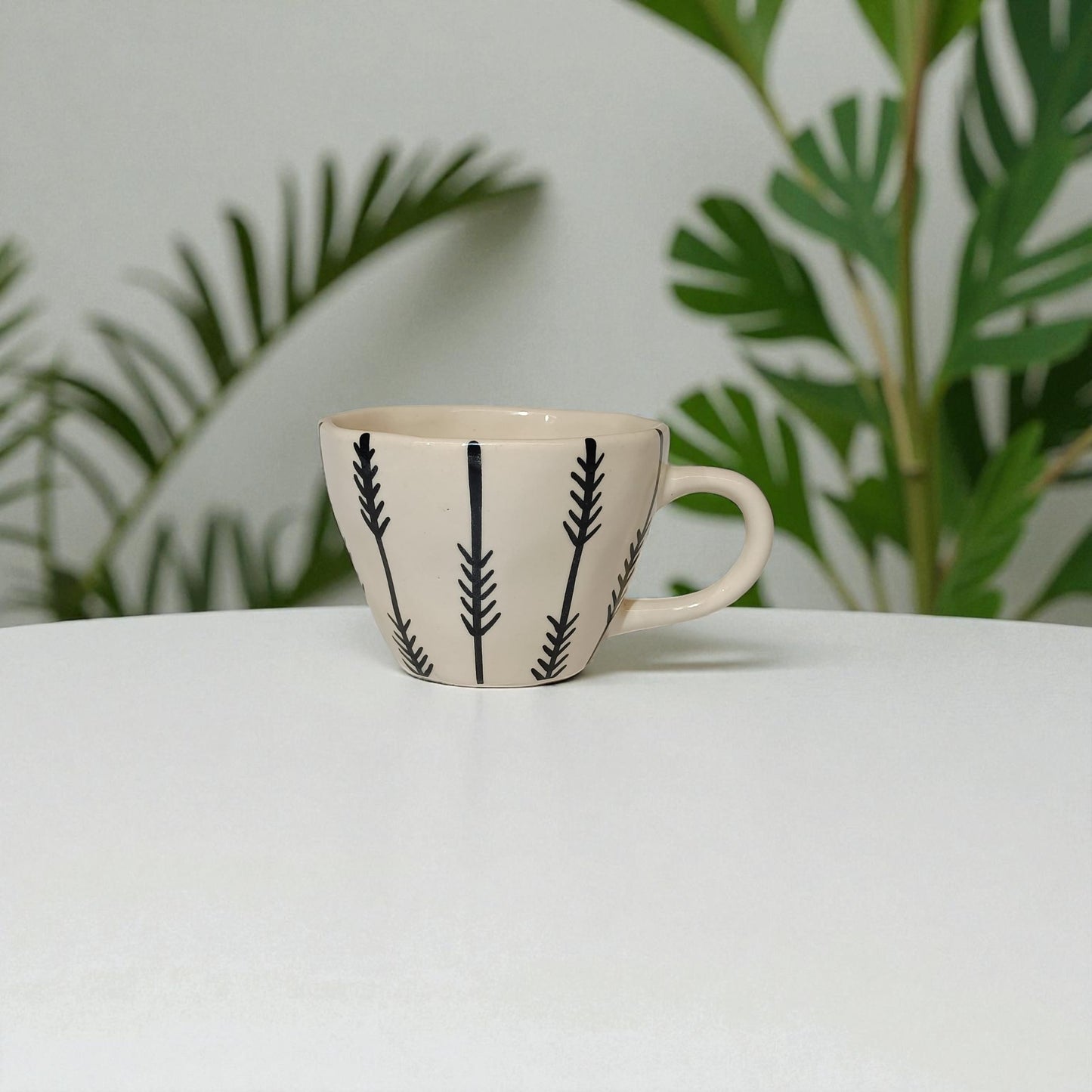 Ceramic mug with black leaf pattern on a white surface with green leaves in the background