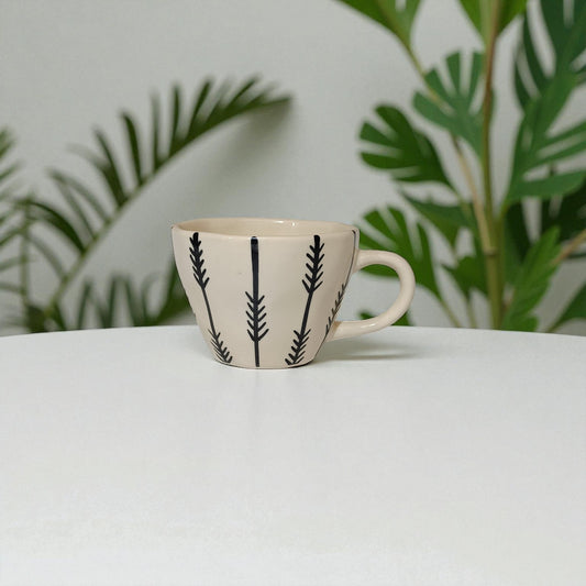 Ceramic mug with black leaf pattern on a white surface with green leaves in the background