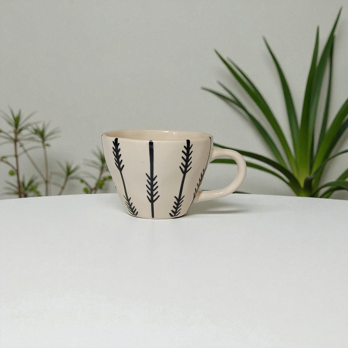 Ceramic mug with black leaf pattern on a white surface with plants in the background