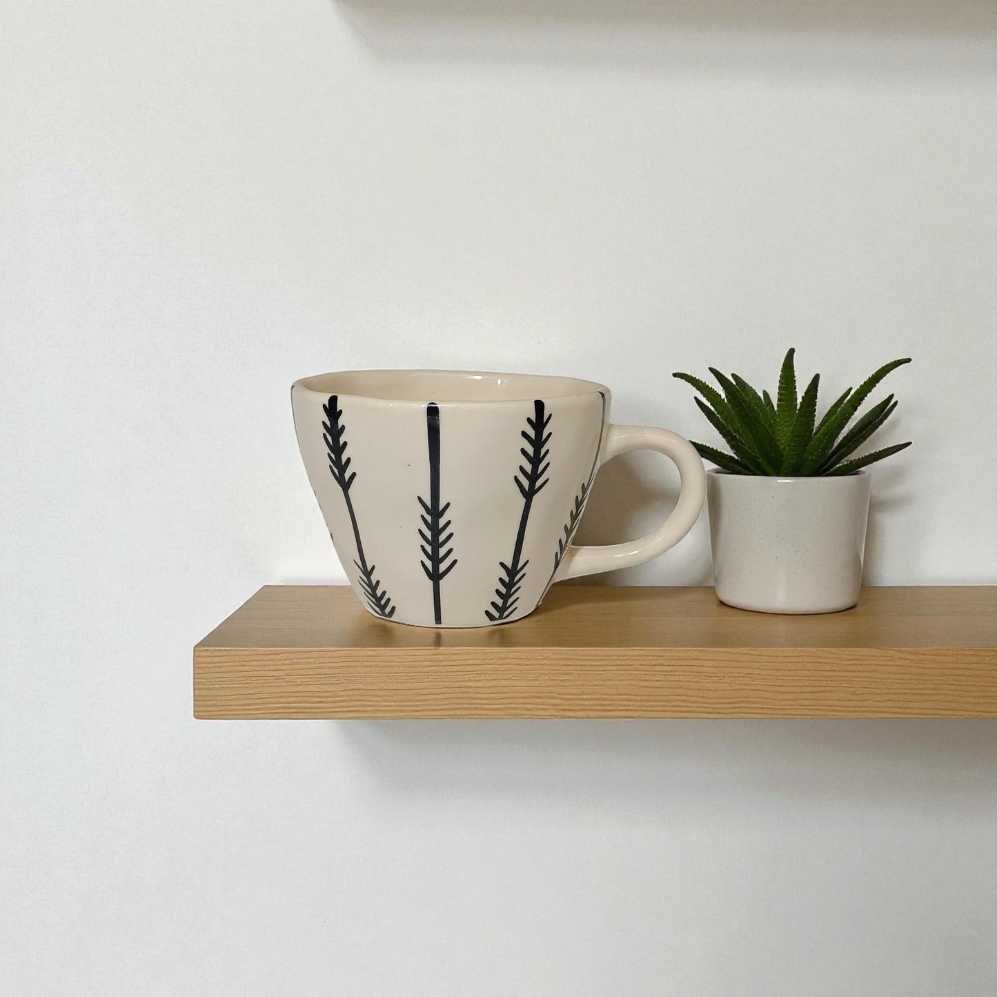Ceramic mug with black leaf pattern on a wooden shelf against a white wall