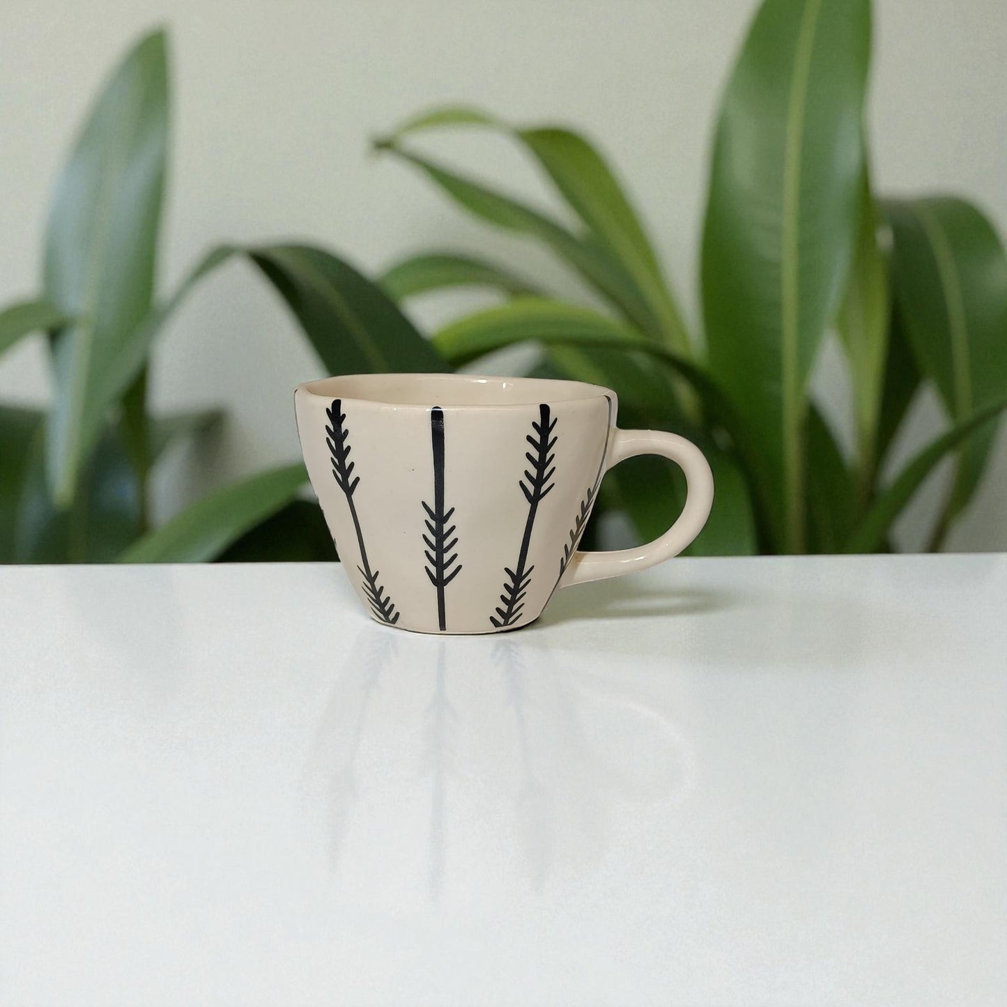 Ceramic mug with black leaf patterns on a white surface with plants in the background