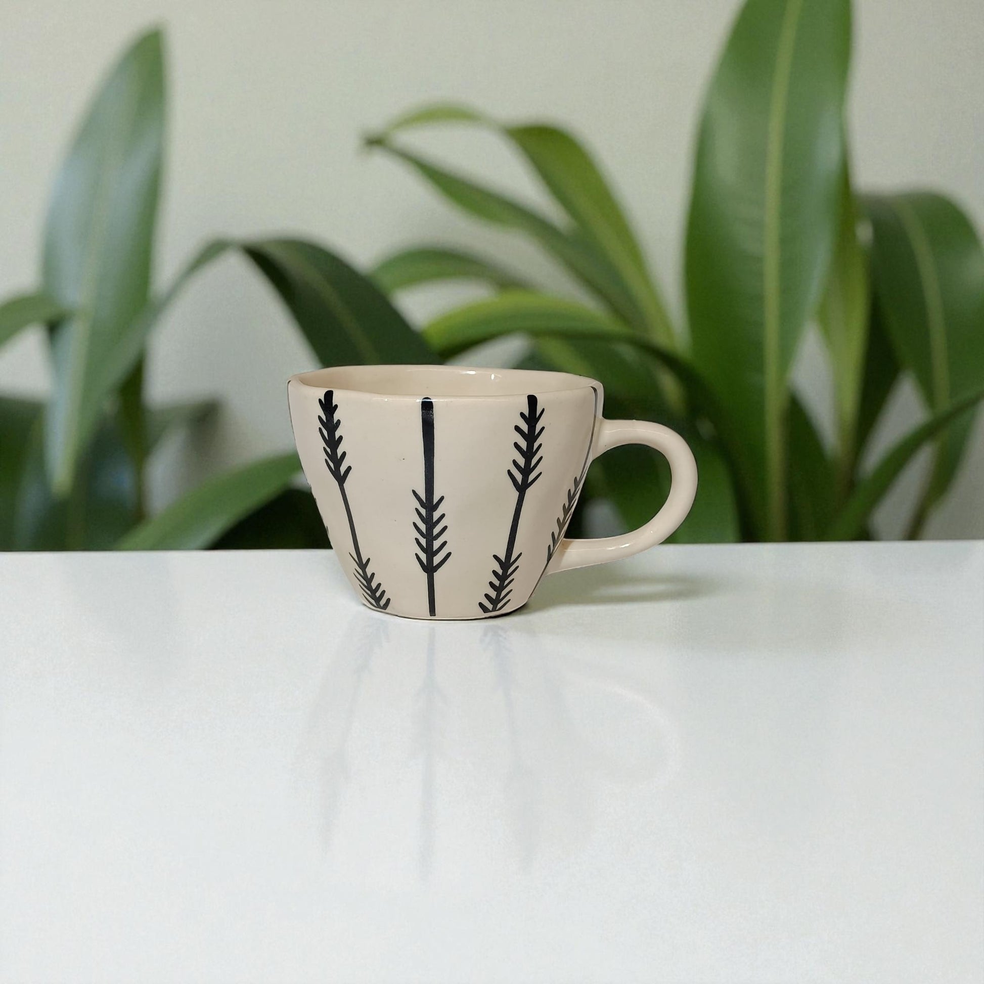 Ceramic mug with black leaf patterns on a white surface with plants in the background