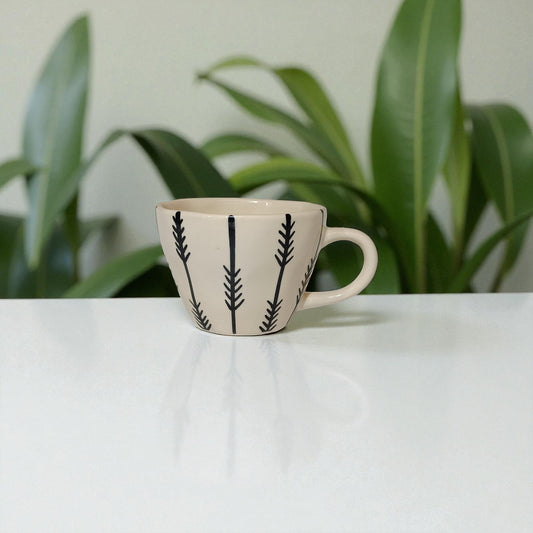 Ceramic mug with black leaf patterns on a white surface with plants in the background