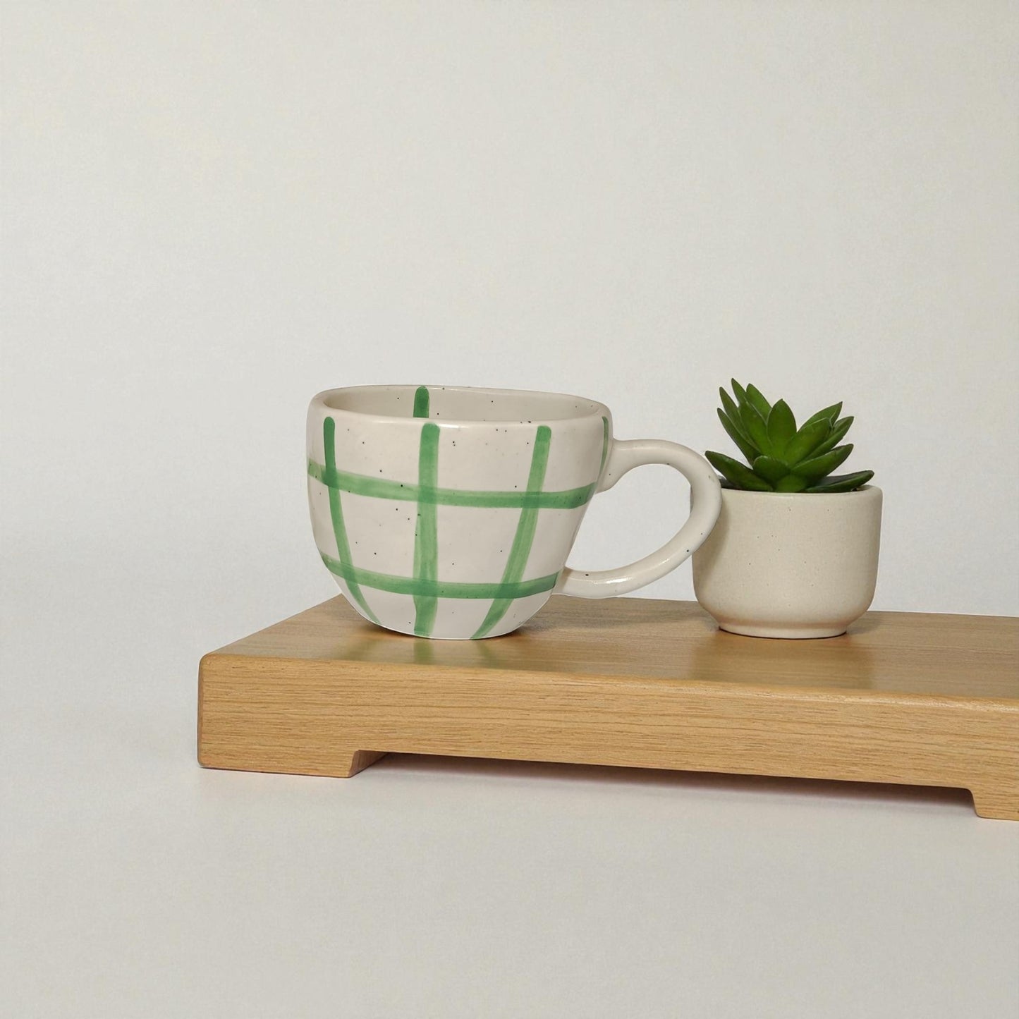 Ceramic mug with green checkered pattern and small plant on a wooden stand against a plain background