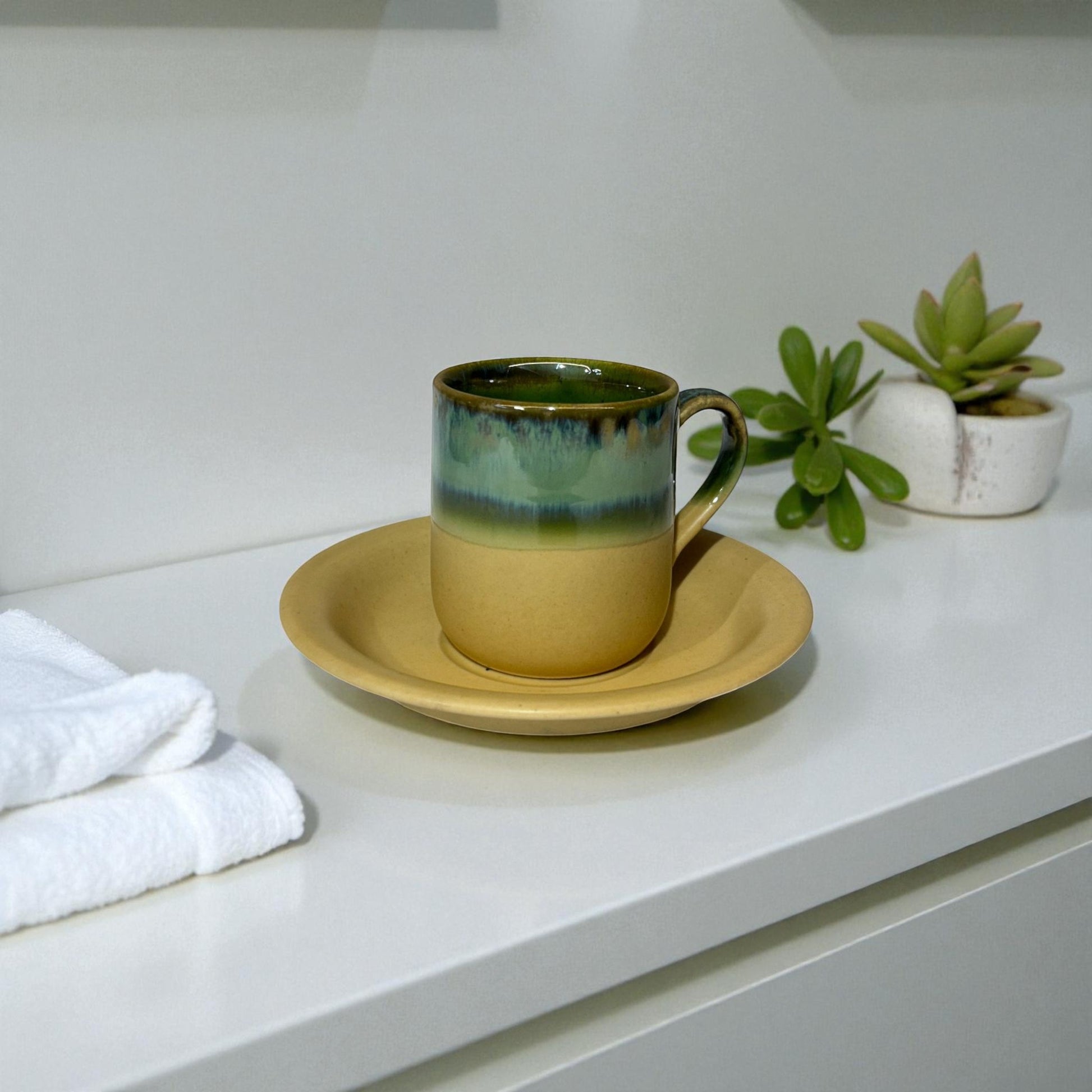 Ceramic mug with green interior and beige exterior on a matching saucer, placed on a white surface with a plant in the background.