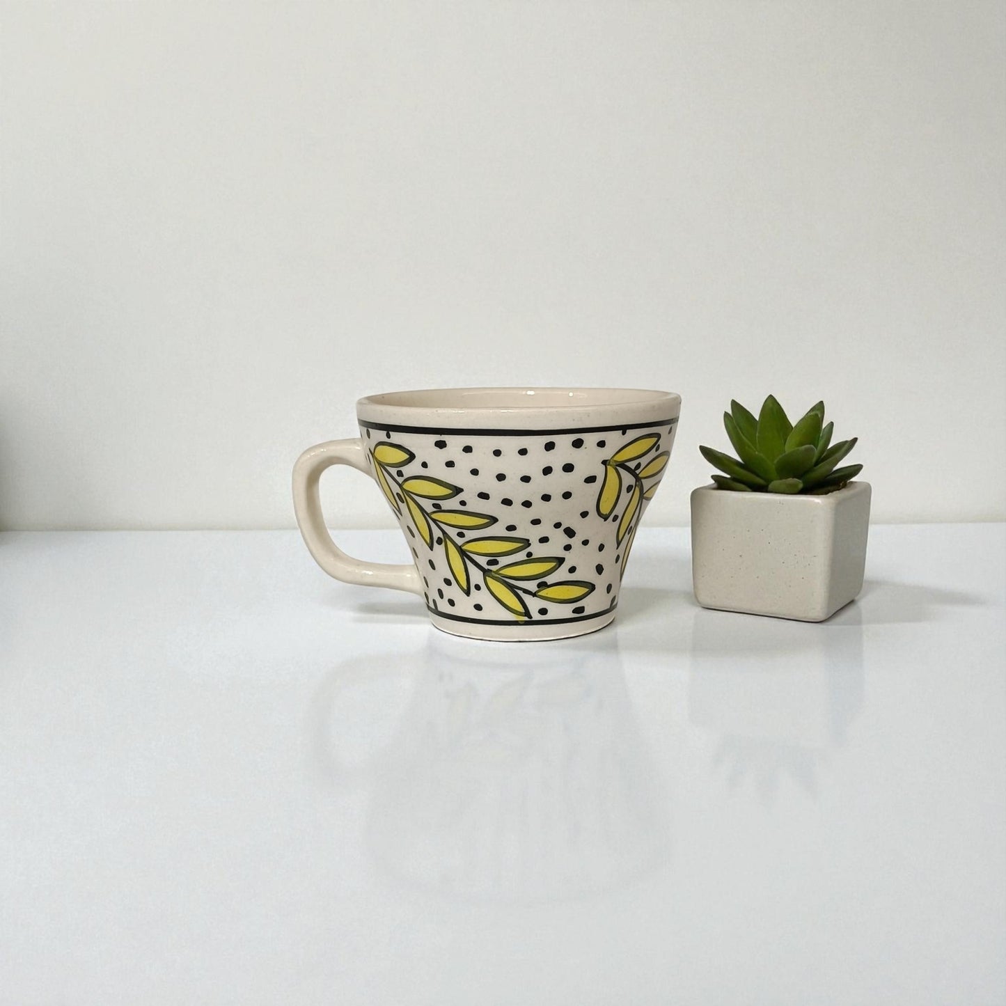 Ceramic mug with leaf pattern and polka dots next to a small potted plant on a white surface.