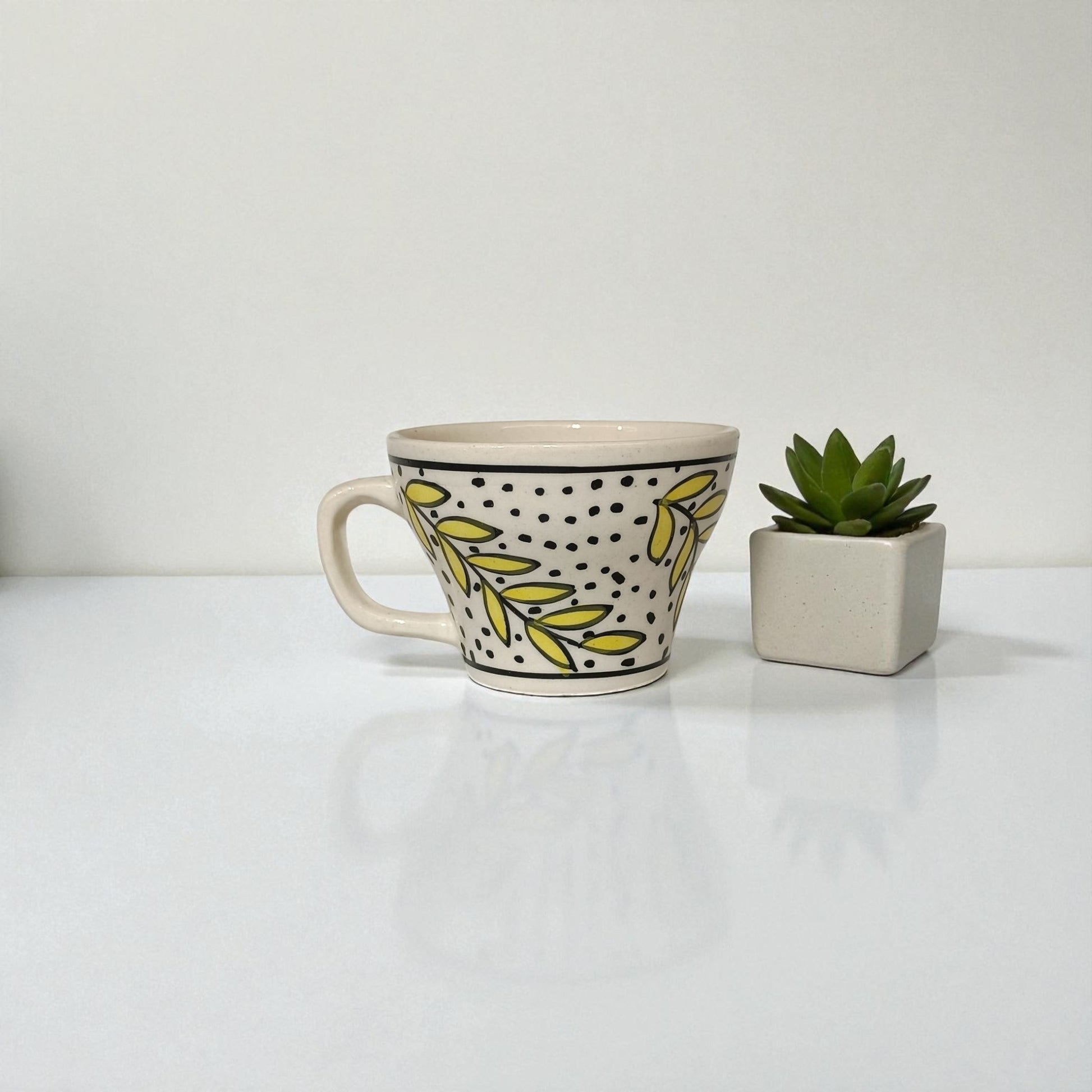 Ceramic mug with leaf pattern and polka dots next to a small potted plant on a white surface.