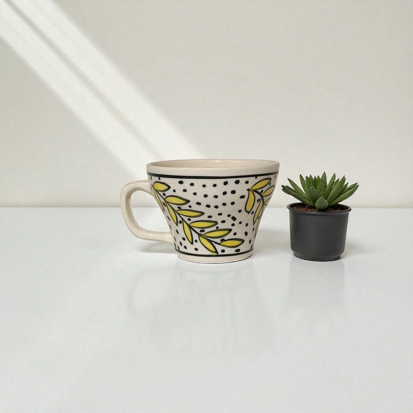 Ceramic mug with leaf pattern next to a small potted plant on a white surface.