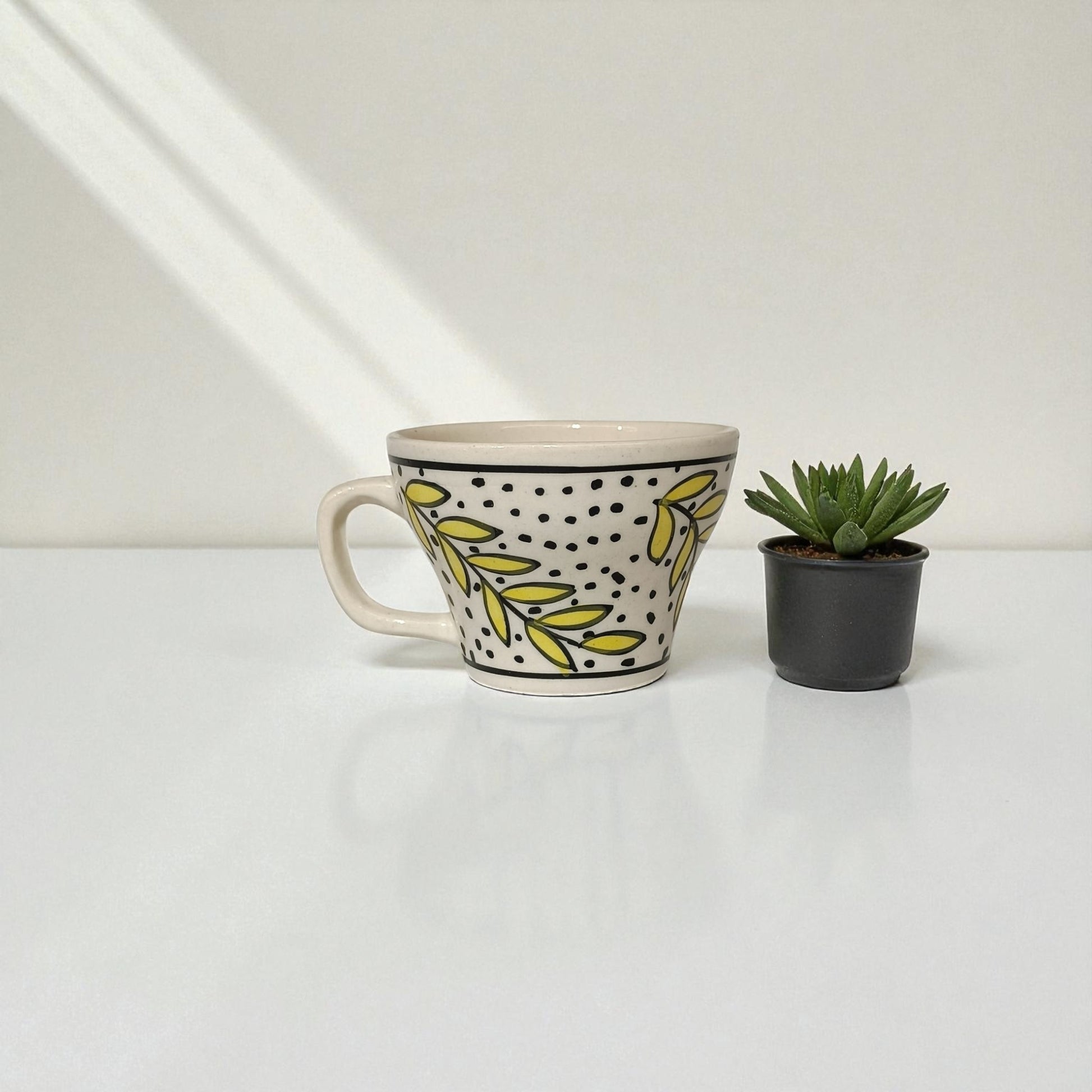 Ceramic mug with leaf pattern next to a small potted plant on a white surface.