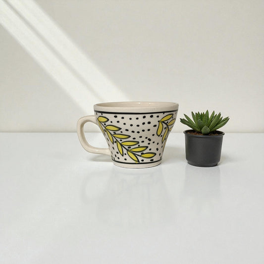 Ceramic mug with leaf pattern next to a small potted plant on a white surface.