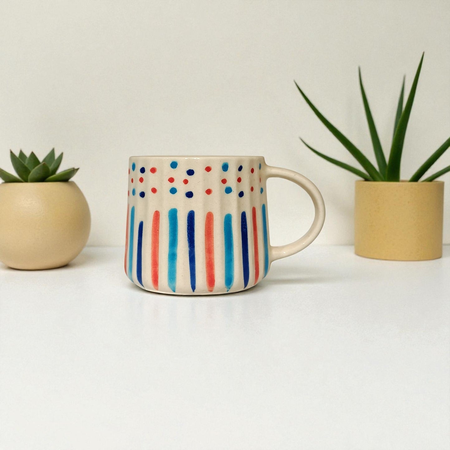 Colorful mug with polka dot and stripe design between two small plants on a light background
