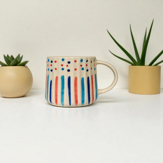 Colorful mug with polka dot and stripe design between two small plants on a light background