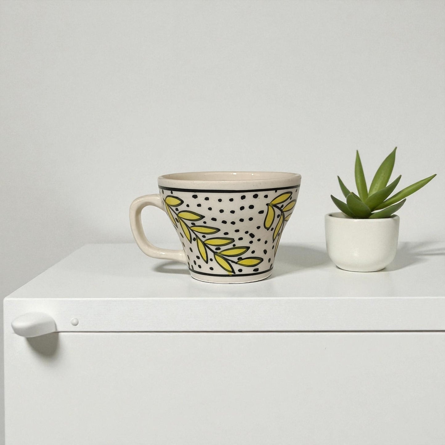 Decorative mug with leaf pattern next to a small potted plant on a white surface.