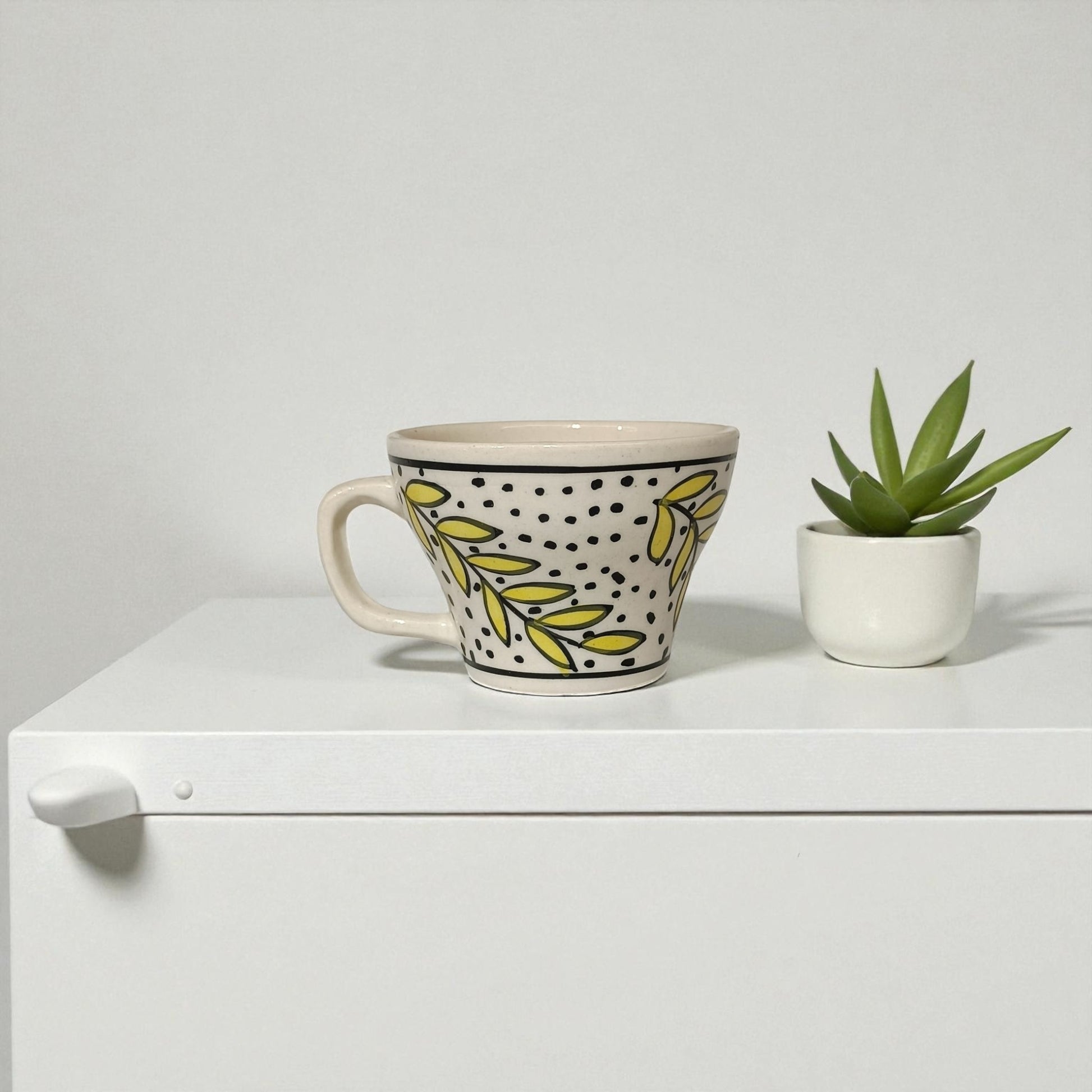 Decorative mug with leaf pattern next to a small potted plant on a white surface.