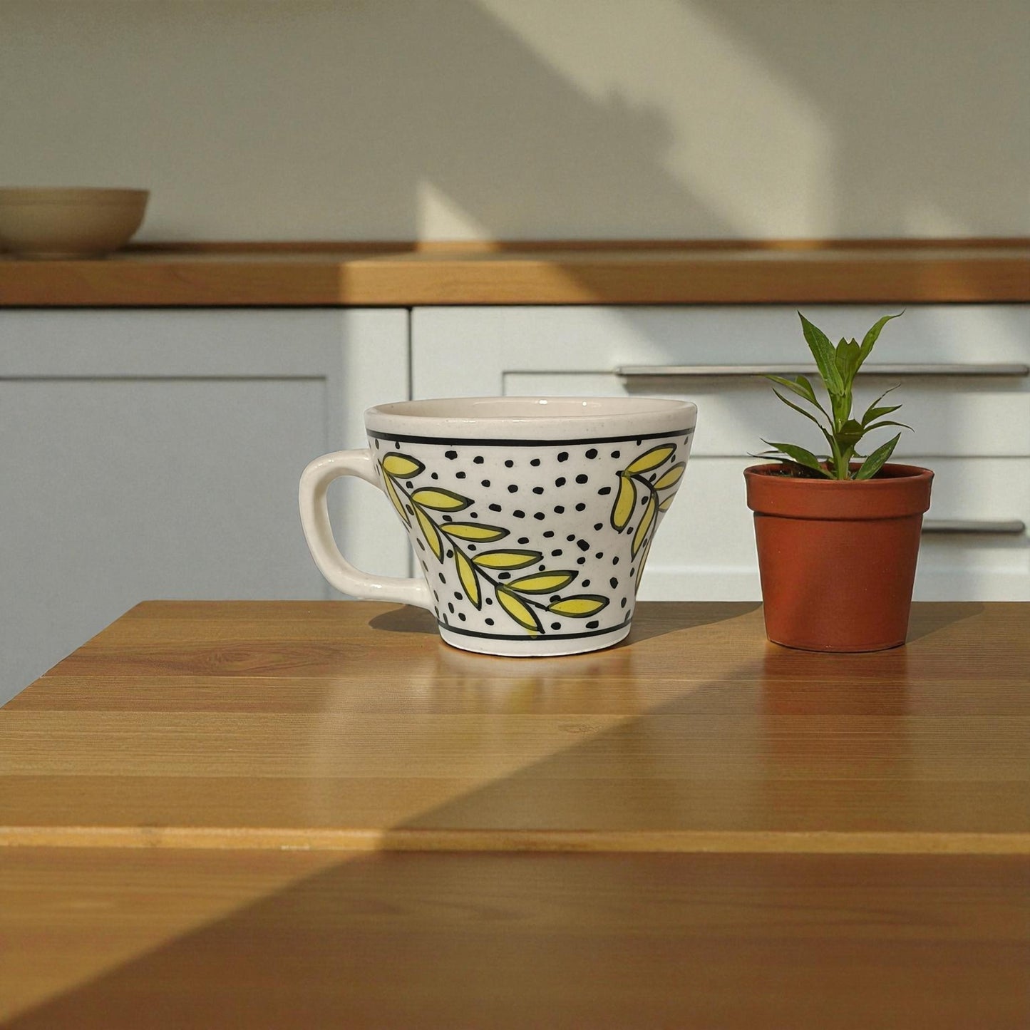 Decorative mug with yellow leaf pattern on a wooden surface next to a potted plant