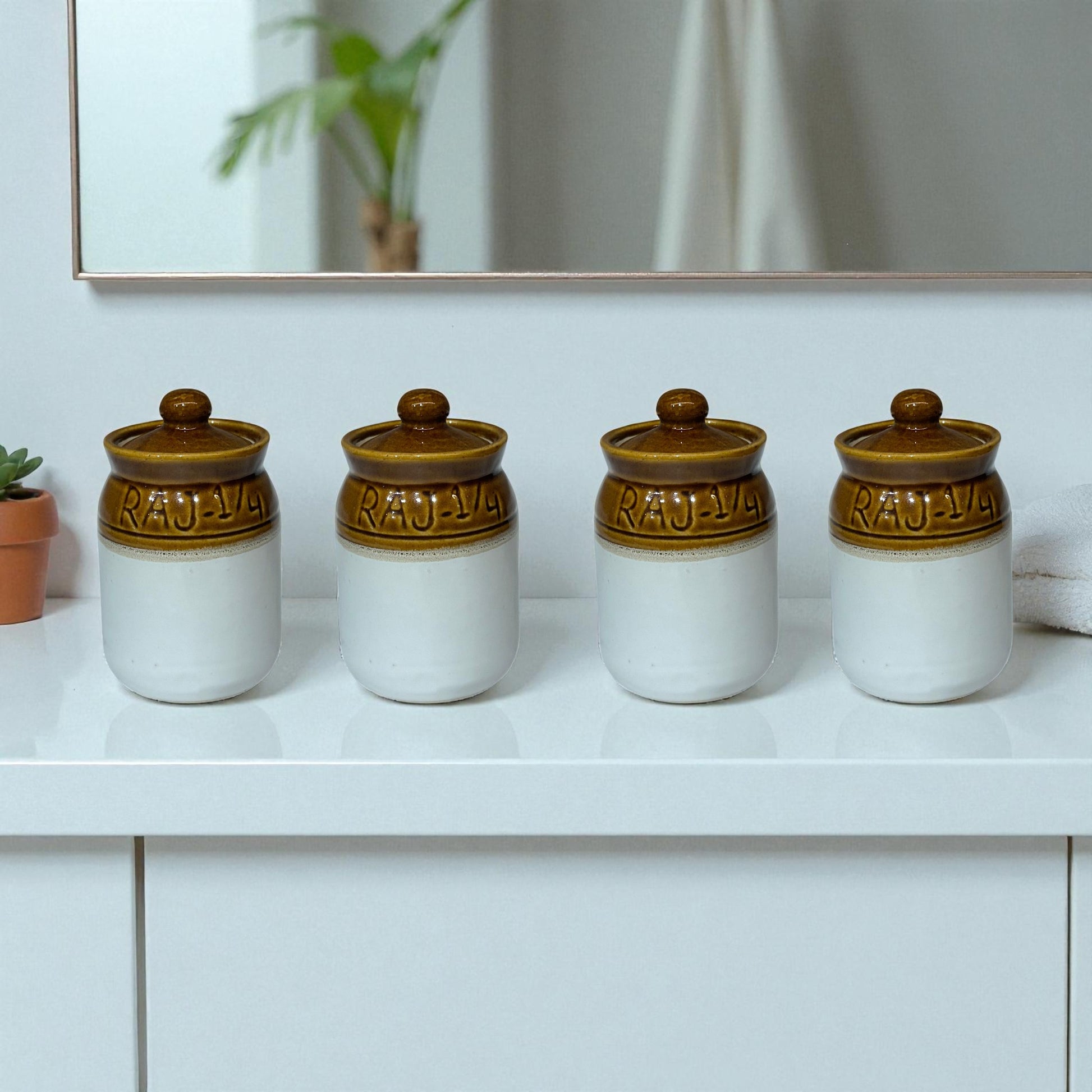 Four ceramic jars with brown lids on a white surface, with a mirror and plant in the background.