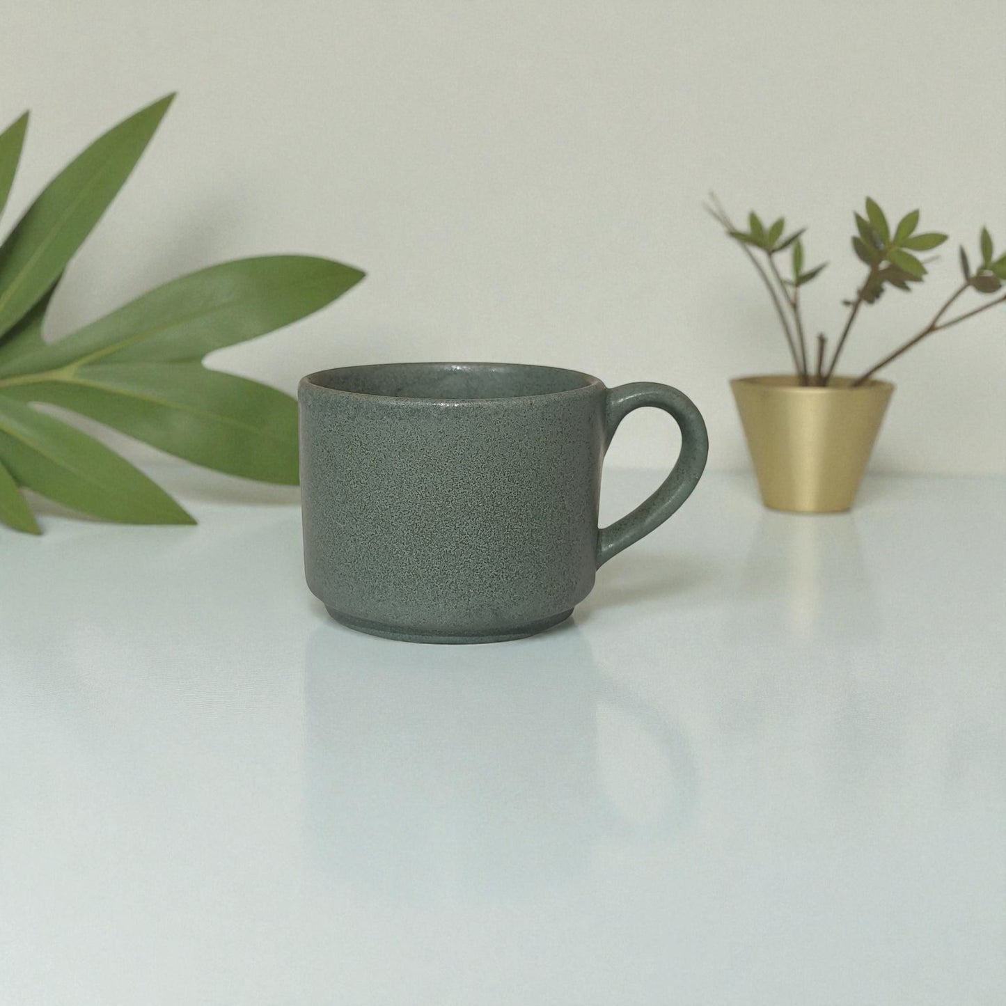 Gray mug on a white surface with green leaves and a gold pot in the background