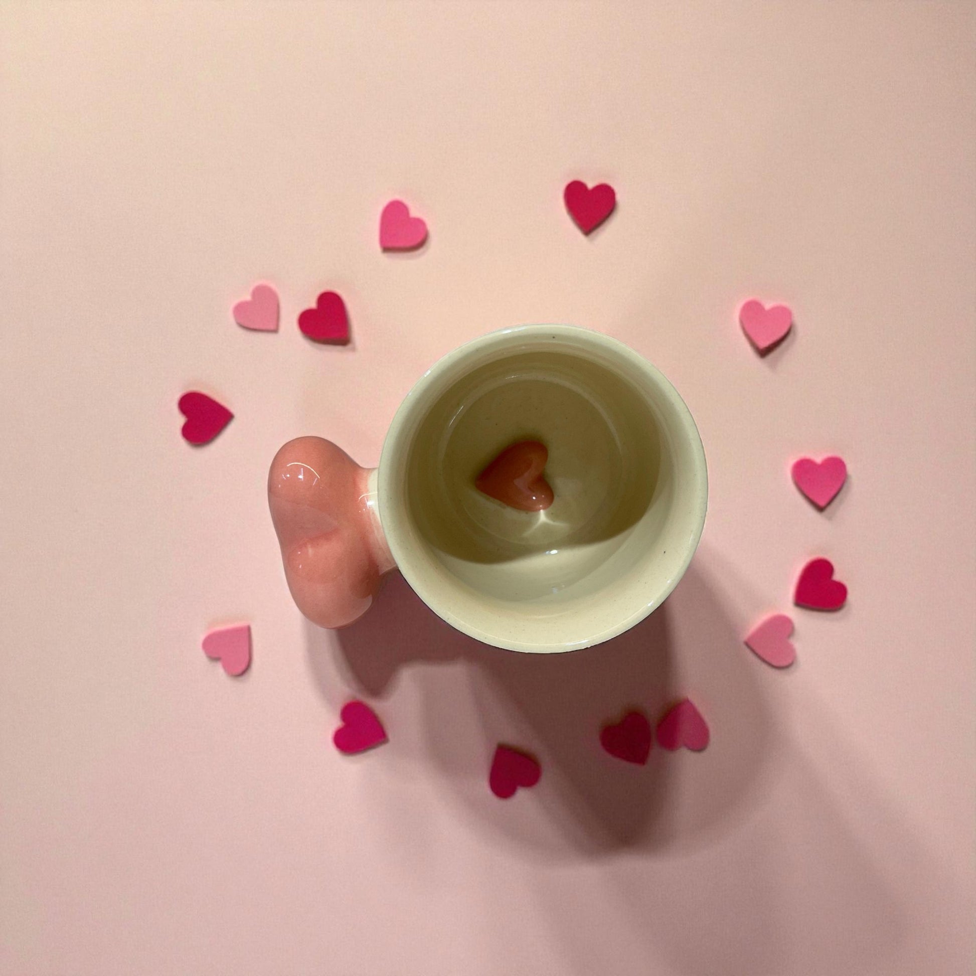 Hand holding a small cup with a heart-shaped object on a pink background with scattered hearts.