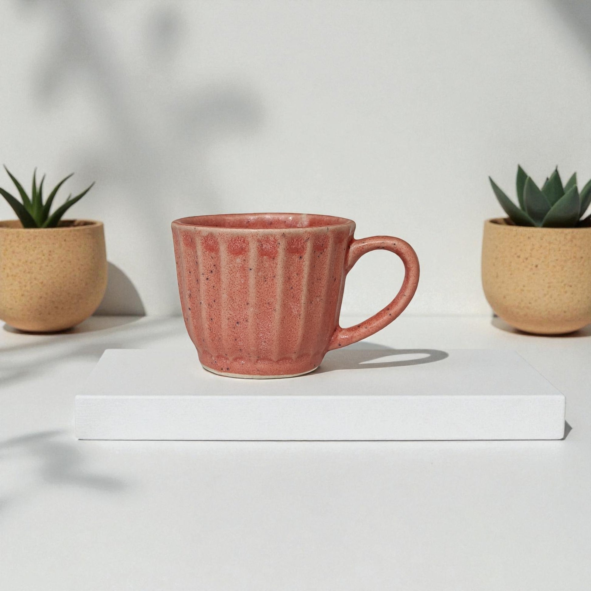 Pink mug on a white surface with two small plants in the background