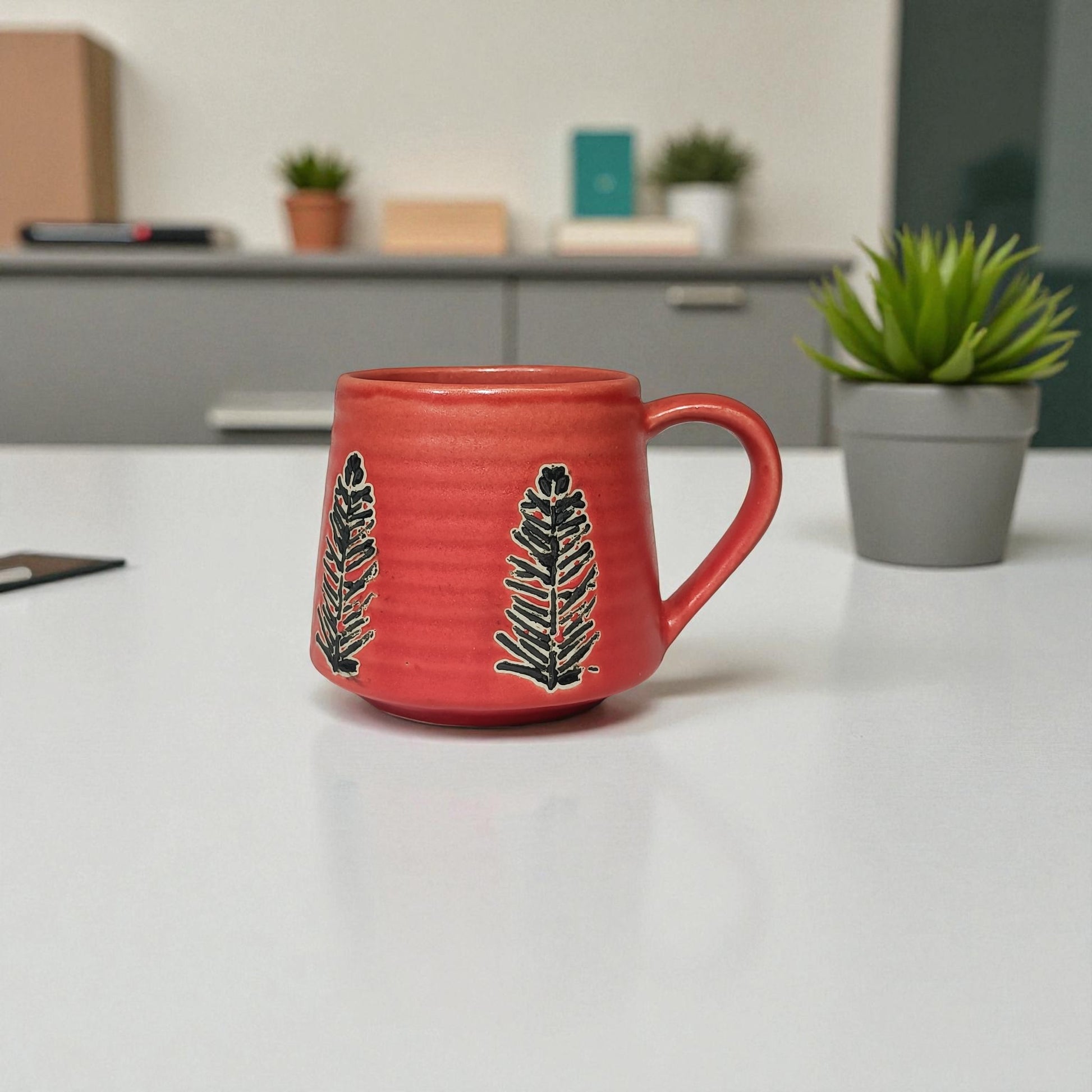 Red mug with black leaf design on a white surface, with a blurred background of a kitchen.