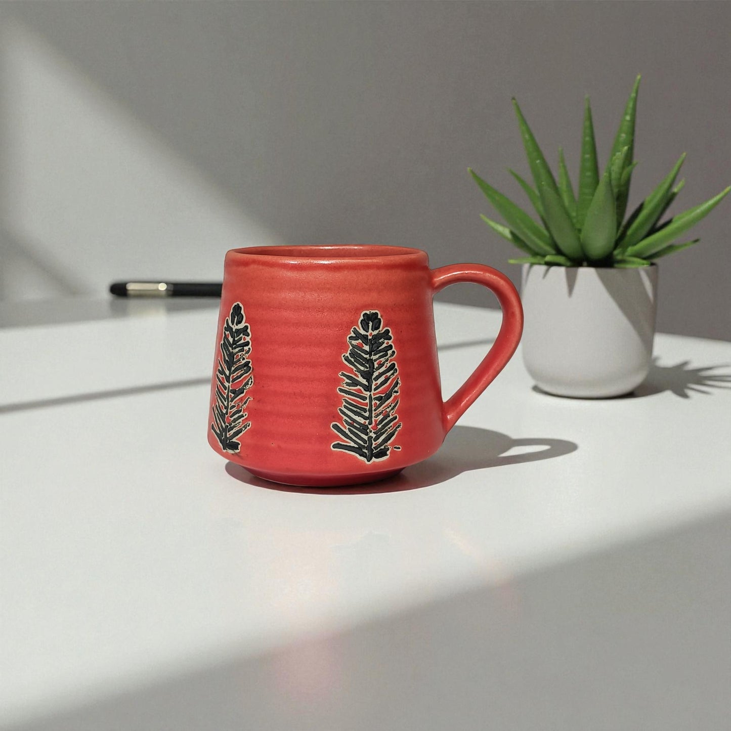 Red mug with leaf design on a white surface next to a green plant