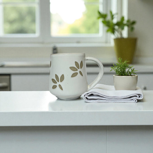 White mug with green leaf pattern on a kitchen counter with a plant and folded towel.